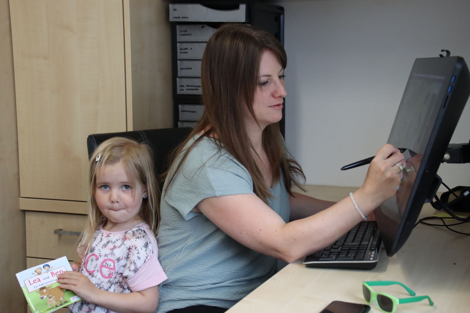 Angela Oberhänsli dessine avec son stylo sur l'écran de son ordinateur, à côté de sa fille qui tient le premier tome de « Léa et Ben ».